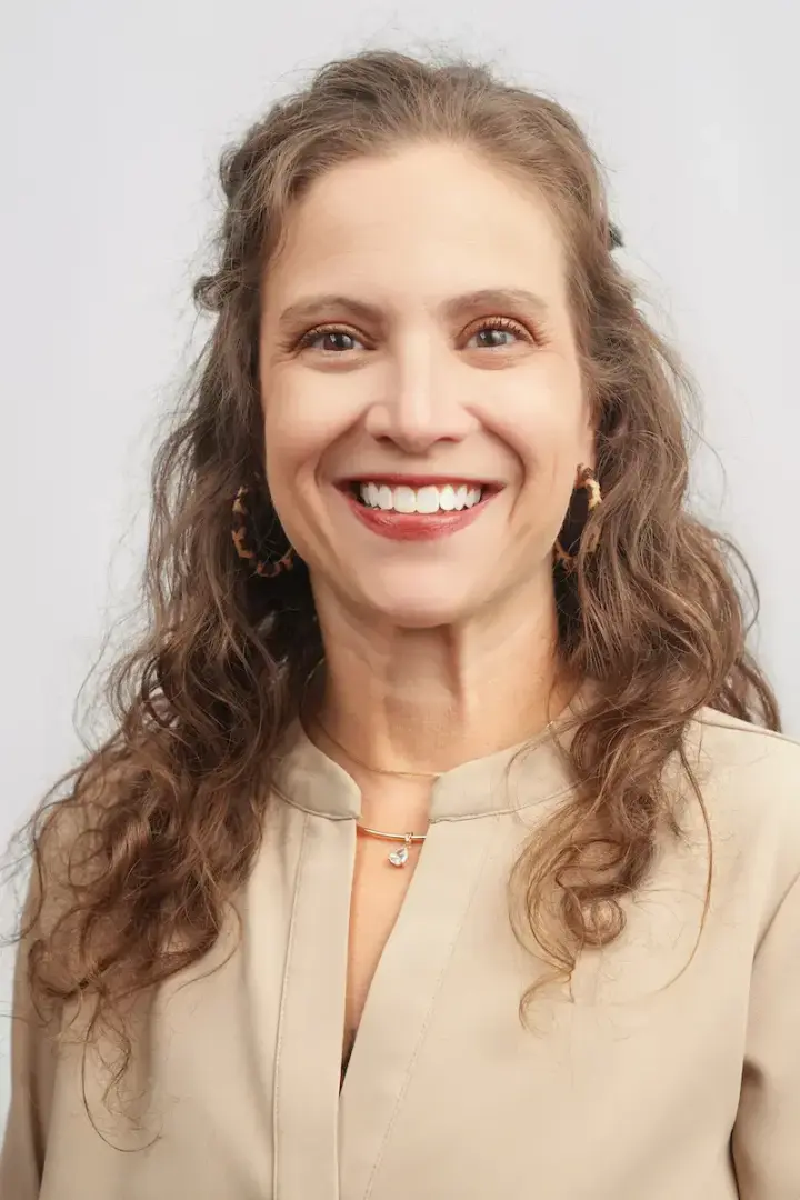 Smiling woman with wavy brown hair in a beige blouse and hoop earrings, facing the camera.